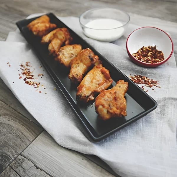 Six crispy chicken wings on a black platter with ranch and chili flakes.