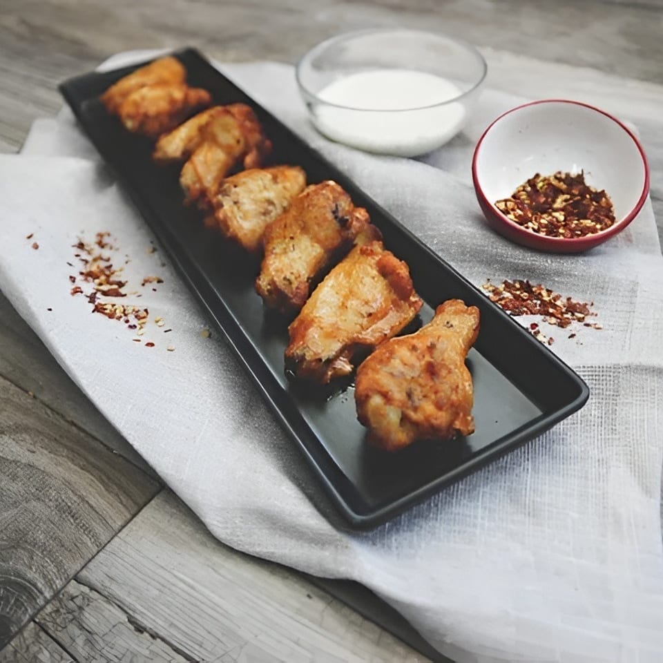 Six crispy chicken wings on a black platter with ranch and chili flakes.