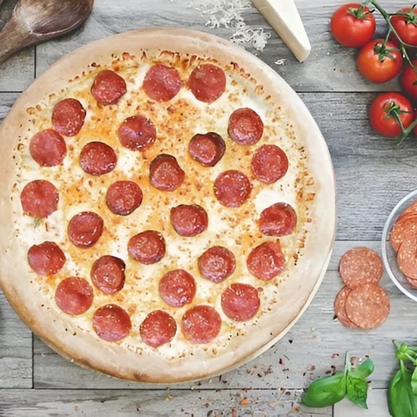 Pepperoni pizza on a wooden table surrounded by tomatoes, basil, extra pepperoni, cheese, and cooking utensils.