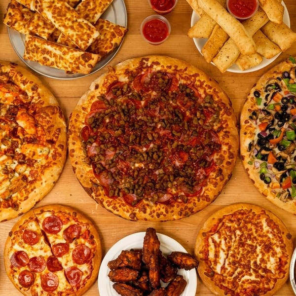 An overhead shot of a wooden table laden with various pizzas, fries, breadsticks, wings, and a cookie.