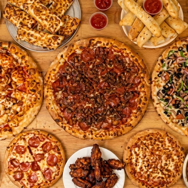 An overhead shot of a wooden table laden with various pizzas, breadsticks, chicken wings, and nuggets.