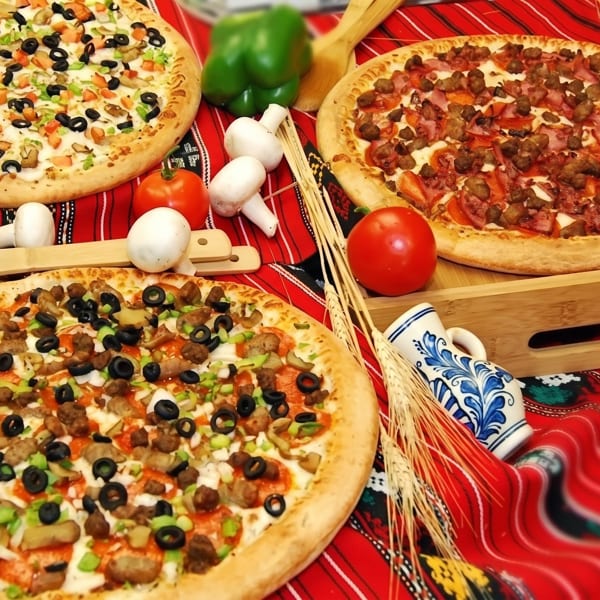 Three pizzas with various toppings, fresh vegetables, and wheat stalks on a red patterned tablecloth.