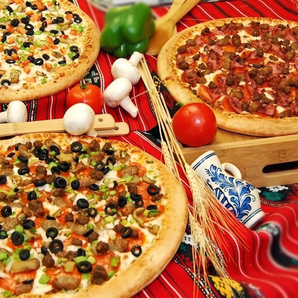 Three pizzas with various toppings, fresh vegetables, and wheat stalks on a red patterned tablecloth.